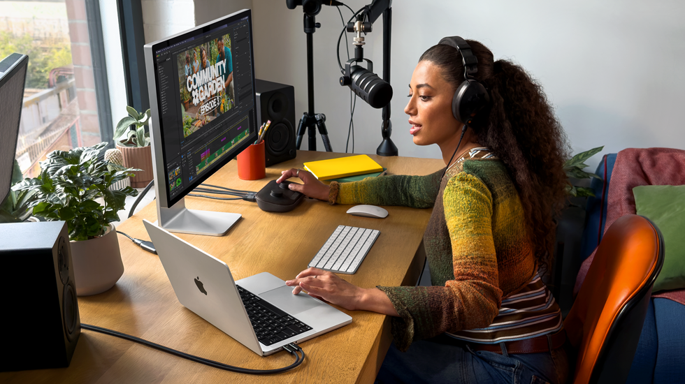 A woman in her home working at her desk with MacBook Pro connected to an external display, wearing headphones and speaking into a microphone