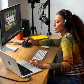 A woman in her home working at her desk with MacBook Pro connected to an external display, wearing headphones and speaking into a microphone
