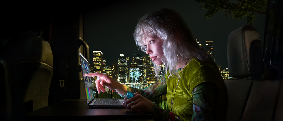 A person in a mobile studio, visible through the open van door, working on a MacBook Pro with PROLED XDR Display, at a remote location near mountains