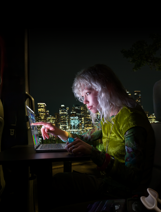 A person in a mobile studio, visible through the open van door, working on a MacBook Pro with PROLED XDR Display, at a remote location near mountains